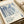 May include: Framed art print featuring abstract blue and white organic flowing patterns with dotted textures, displayed on white bedding next to a beige pillow. The poster shows Japanese text at bottom with "Tokyo, Japan 1984" and color swatches. The artwork has fluid, wave-like forms in various shades of blue against cream background, presented in a thin light wood frame.