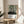 May include: Framed art print featuring abstract forest scene with black vertical tree trunks, green circular shapes, and dotted ground pattern on cream background. Text reads "YAYOI KUSAMA" with Japanese characters and "Tokyo, Japan 1984" plus green color palette swatches. Light wood frame sits on round wooden side table next to beige upholstered chair with white pillow. Plant leaves visible in corner, natural window light creates soft shadows on white wall.