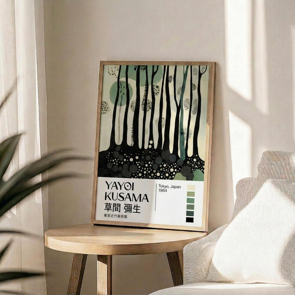 May include: Framed art print featuring abstract forest scene with black vertical tree trunks, green circular shapes, and dotted ground pattern on cream background. Text reads "YAYOI KUSAMA" with Japanese characters and "Tokyo, Japan 1984" plus green color palette swatches. Light wood frame sits on round wooden side table next to beige upholstered chair with white pillow. Plant leaves visible in corner, natural window light creates soft shadows on white wall.