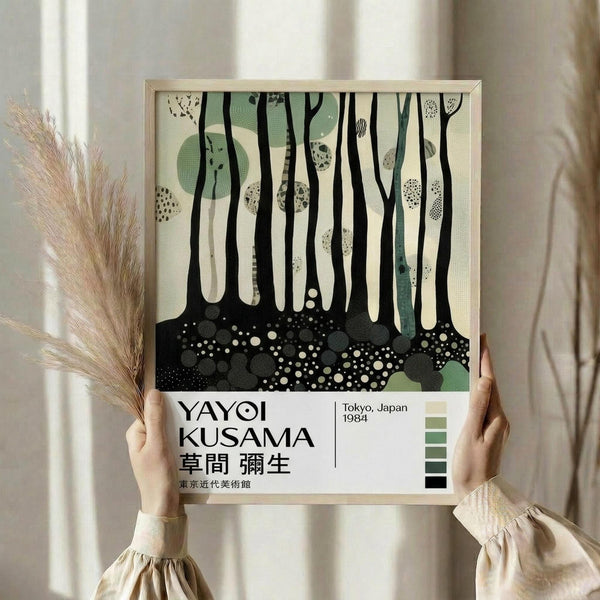 May include: Person holding framed poster in natural light setting with dried pampas grass nearby. The artwork features abstract forest scene with tall black tree trunks, sage green circular shapes, white dotted patterns, and speckled ground in cream and black tones. Exhibition-style text below reads "YAYOI KUSAMA" with Japanese characters, "Tokyo, Japan 1984" and includes small color palette squares in greens and black.