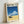 May include: Framed poster print leaning against white wall showing retro-style artwork with blue ocean waves, fluffy white clouds, yellow sandy beach, and dark vintage car. Features "HIROSHI NAGAI" text at top with Japanese characters below. Light wood frame surrounds the vibrant coastal scene with clear blue sky gradient.
