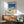May include: Mid-century modern living room scene with framed poster on white wall showing vintage car on sandy beach beneath blue sky and white clouds. Wooden credenza below holds ceramic table lamp. Teal upholstered armchair with wooden frame sits nearby alongside small round coffee table. Natural sunlight creates dramatic shadows across the minimalist interior space.