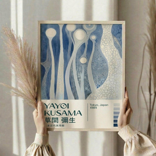May include: Person in beige clothing holding framed art poster featuring abstract blue and cream organic shapes with dotted patterns. Dried pampas grass visible to left. Soft natural lighting against neutral curtained background. Poster shows flowing, cellular-like forms in various shades of blue with white circular elements and intricate stippled textures throughout the composition.