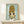 May include: Framed artwork featuring an anime-style character with large brown beehive hair and oversized dark eyes, wearing a yellow top against a soft blue background. The piece sits on a light wooden shelf next to a small white wrapped gift with greenery. Natural lighting creates gentle shadows on the cream-colored wall behind.