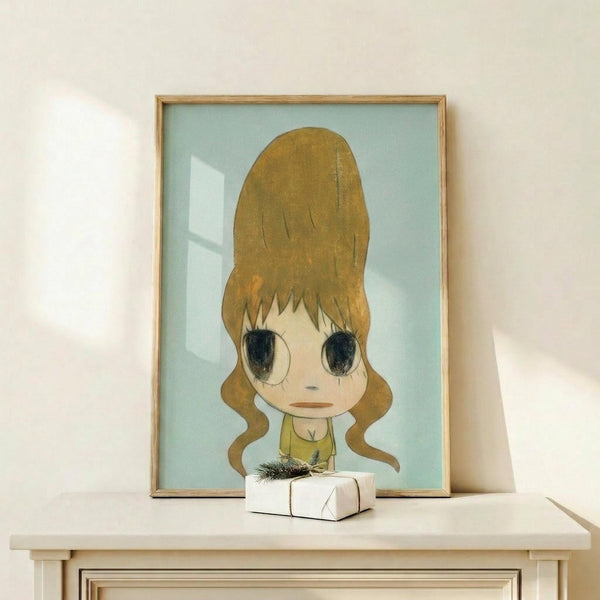 May include: Framed artwork featuring an anime-style character with large brown beehive hair and oversized dark eyes, wearing a yellow top against a soft blue background. The piece sits on a light wooden shelf next to a small white wrapped gift with greenery. Natural lighting creates gentle shadows on the cream-colored wall behind.