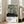 May include: Framed art print leaning against white wall next to beige sofa in bright living room. Abstract forest scene with black tree trunks, dotted ground, and green circular shapes on cream background. White exhibition-style text panel below artwork. Natural wood frame, approximately 2-3 feet tall, positioned on light wooden floor near large windows.
