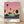 May include: Framed poster print leaning against white wall on wooden floor. Artwork shows retro-style illustration of turquoise mid-century modern building with illuminated windows, surrounded by tall palm trees silhouetted against gradient sunset sky transitioning from pink to peach. Dark road runs across bottom. Text reads "HIROSHI NAGAI" and "TOKYO 1973" with color palette dots. Natural light casts soft shadows on wall.