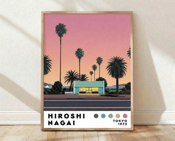 May include: Framed poster print leaning against white wall on wooden floor. Artwork shows retro-style illustration of turquoise mid-century modern building with illuminated windows, surrounded by tall palm trees silhouetted against gradient sunset sky transitioning from pink to peach. Dark road runs across bottom. Text reads "HIROSHI NAGAI" and "TOKYO 1973" with color palette dots. Natural light casts soft shadows on wall.