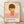 May include: Framed poster print leaning against white wall on light wood floor. Artwork shows minimalist illustration of child-like figure with brown bob haircut, closed eyes, wearing orange dress with white daisy flower. Figure stands against peachy background with pink ground. Text reads "YOSHITOMO NARA" with Japanese characters and exhibition dates below. Natural wood frame with white mat.