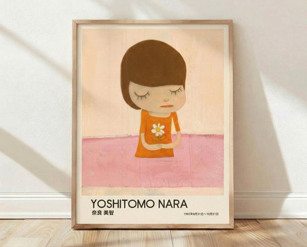 May include: Framed poster print leaning against white wall on light wood floor. Artwork shows minimalist illustration of child-like figure with brown bob haircut, closed eyes, wearing orange dress with white daisy flower. Figure stands against peachy background with pink ground. Text reads "YOSHITOMO NARA" with Japanese characters and exhibition dates below. Natural wood frame with white mat.
