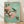 May include: Framed Japanese woodblock print featuring a small green and white bird perched on a blooming cherry blossom branch with delicate pink flowers against a soft turquoise background. The artwork displays traditional Japanese calligraphy and a red seal stamp, presented in a light wood frame leaning against white walls on light wooden flooring with natural shadows.