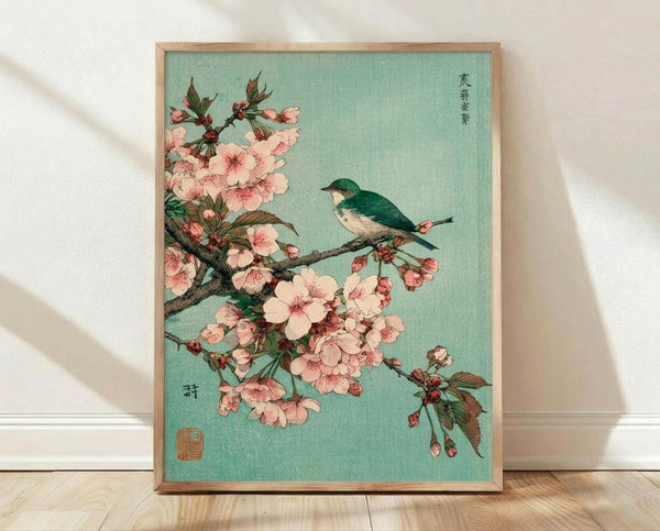 May include: Framed Japanese woodblock print featuring a small green and white bird perched on a blooming cherry blossom branch with delicate pink flowers against a soft turquoise background. The artwork displays traditional Japanese calligraphy and a red seal stamp, presented in a light wood frame leaning against white walls on light wooden flooring with natural shadows.