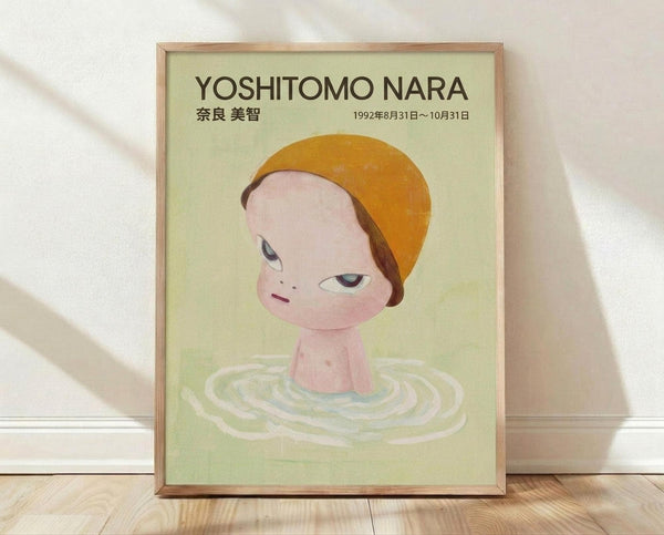 May include: Framed poster featuring a childlike figure with orange hair and pink skin emerging from rippling water against a pale green background. The artwork shows Japanese text and exhibition dates from 1992. The wooden frame leans against a white wall on light wooden flooring, with soft natural lighting creating gentle shadows.