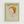 Framed poster featuring a stylized illustration of a child-like figure with an oversized head, orange bowl-cut hair, and large eyes, emerging from concentric ripples in pale green water. The artwork has a soft, dreamy quality with muted colors on a light green background. Text at the top reads "YOSHITOMO NARA" with Japanese characters and exhibition dates from 1992. The poster is displayed in a natural wood frame against a white wall.