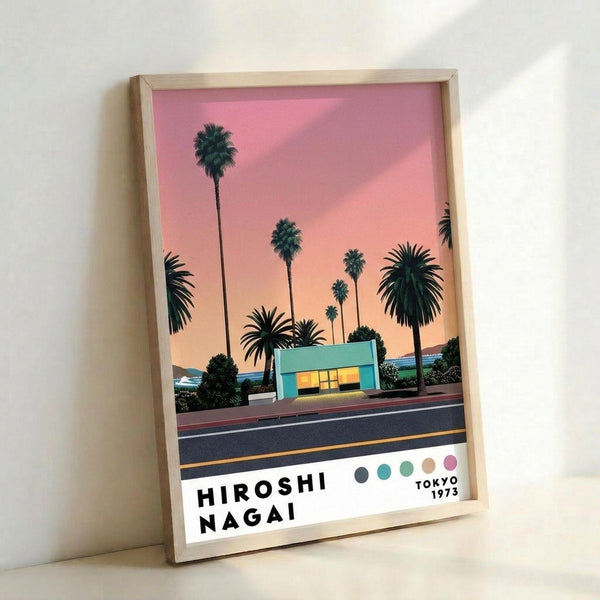 A framed poster artwork featuring palm trees silhouetted against a pink and orange sunset sky, with a turquoise modern building and road in the foreground. The print has "HIROSHI NAGAI" and "TOKYO 1973" text at the bottom along with a row of colored dots. The artwork is displayed in a light wood frame leaning against a white wall.