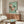 May include: Japanese woodblock print artwork featuring pink cherry blossoms and a small green bird on branches against a soft mint green background, displayed in thin wooden frame above mid-century modern walnut credenza with white ceramic table lamp, cream upholstered round chair, and oval glass coffee table in minimalist living room with white walls and natural lighting.