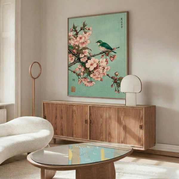 May include: Japanese woodblock print artwork featuring pink cherry blossoms and a small green bird on branches against a soft mint green background, displayed in thin wooden frame above mid-century modern walnut credenza with white ceramic table lamp, cream upholstered round chair, and oval glass coffee table in minimalist living room with white walls and natural lighting.