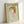 May include: Framed vintage-style exhibition poster featuring a stylized illustration of a child's head and shoulders emerging from rippling water. The figure has pale skin, an orange bowl-cut hairstyle, and large eyes with a contemplative expression. Set against a soft green background with Japanese text at the top and exhibition dates. Displayed in a light wooden frame leaning against a white wall, creating subtle shadows. The poster has a dreamy, ethereal quality with muted pastel tones throu