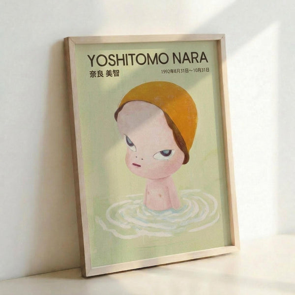 May include: Framed vintage-style exhibition poster featuring a stylized illustration of a child's head and shoulders emerging from rippling water. The figure has pale skin, an orange bowl-cut hairstyle, and large eyes with a contemplative expression. Set against a soft green background with Japanese text at the top and exhibition dates. Displayed in a light wooden frame leaning against a white wall, creating subtle shadows. The poster has a dreamy, ethereal quality with muted pastel tones throu
