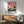 May include: Framed poster print on white wall above wood sideboard, featuring palm trees against pink sunset sky with modernist building. Mid-century modern living room with sage green upholstered chair, round coffee table, and ceramic table lamp. Warm natural lighting creates geometric shadows on wall.