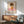 May include: Framed poster print featuring minimalist illustration of child with brown bob haircut, closed eyes, wearing orange top with white flower, standing on pink surface against beige background. Text reads "YOSHITOMO NARA" below artwork. Displayed above wooden sideboard with white ceramic lamp, next to sage green mid-century modern armchair with wooden frame in bright, naturally-lit room with white walls.