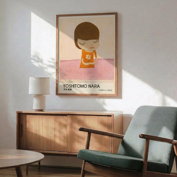 May include: Framed poster print featuring minimalist illustration of child with brown bob haircut, closed eyes, wearing orange top with white flower, standing on pink surface against beige background. Text reads "YOSHITOMO NARA" below artwork. Displayed above wooden sideboard with white ceramic lamp, next to sage green mid-century modern armchair with wooden frame in bright, naturally-lit room with white walls.