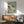 May include: Japanese woodblock print style artwork showing pink cherry blossoms on a branch with a small blue and white bird perched among the flowers, set against a soft mint green background. The framed print hangs above a mid-century modern wooden sideboard next to a sage green upholstered armchair with wooden frame. Natural sunlight creates geometric shadows on the white wall, and a white ceramic table lamp sits on the credenza.