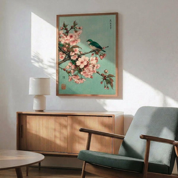 May include: Japanese woodblock print style artwork showing pink cherry blossoms on a branch with a small blue and white bird perched among the flowers, set against a soft mint green background. The framed print hangs above a mid-century modern wooden sideboard next to a sage green upholstered armchair with wooden frame. Natural sunlight creates geometric shadows on the white wall, and a white ceramic table lamp sits on the credenza.