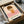 May include: Framed art print featuring a minimalist illustration of a young girl with brown bob haircut and closed eyes, wearing an orange top with white flower design, sitting on pink checkered surface against cream background. Black frame sits in opened cardboard shipping box with white tissue paper padding.