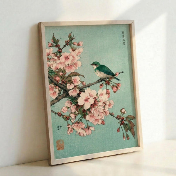 May include: Framed vintage Japanese woodblock print reproduction leaning against white wall. Features small teal and white bird perched on flowering cherry blossom branch with delicate pink petals against soft mint green background. Light wood frame, vertical orientation, positioned on light surface with natural lighting creating subtle shadows.