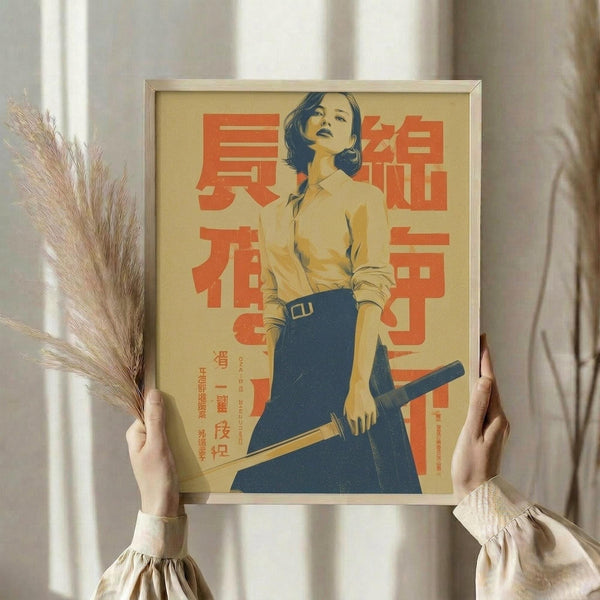 May include: Hands holding a framed vintage-style poster featuring a woman with dark hair in a yellow shirt and blue skirt holding a katana sword, with bold red Japanese text on a cream background. Golden frame, dried wheat stems visible to the left, soft natural lighting from window curtains in background.