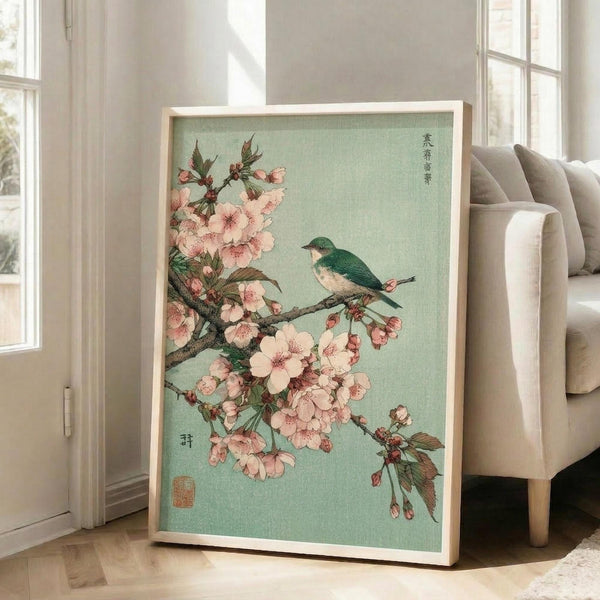 May include: Large framed vintage Japanese woodblock print featuring pink cherry blossoms and a teal bird perched on flowering branches against sage green background, displayed on light wooden floors next to beige upholstered chair in bright sunlit room with white walls and windows.