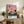 May include: Framed poster with pink-to-orange gradient sunset sky, silhouetted palm trees, and turquoise modernist building along a dark road. Text reads "HIROSHI NAGAI" and "TOKYO 1973" with colored dots. Displayed on rustic wooden console with fern plant, ceramic vase, books, and beige armchair in neutral-toned living room setting.