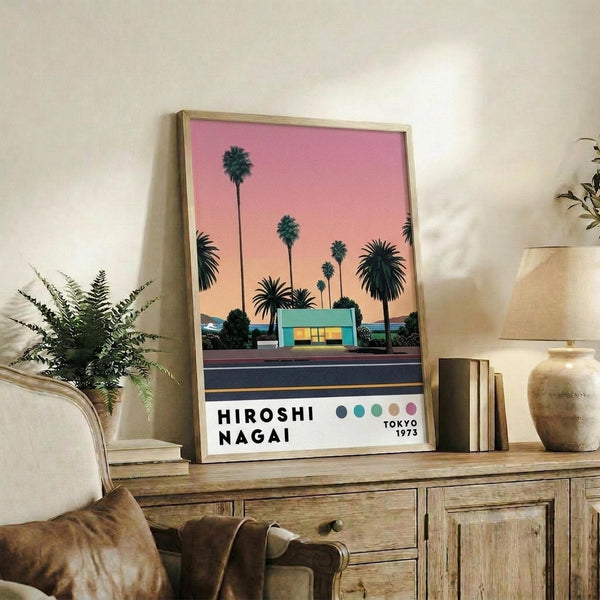 May include: Framed poster with pink-to-orange gradient sunset sky, silhouetted palm trees, and turquoise modernist building along a dark road. Text reads "HIROSHI NAGAI" and "TOKYO 1973" with colored dots. Displayed on rustic wooden console with fern plant, ceramic vase, books, and beige armchair in neutral-toned living room setting.