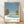 May include: Framed poster print leaning against white textured wall on light wood floor. Artwork shows retro-style illustration of turquoise swimming pool surrounded by white concrete, two large palm trees, and flowering bushes under blue sky. Text reads "HIROSHI NAGAI" and "TOKYO 1973" with colored dots in blue, coral, and olive green. Natural wood frame, soft shadows from window light.