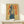 May include: Framed vintage-style Coca-Cola poster featuring a woman in casual clothing holding a Coke bottle, with bold red Japanese text on yellow background. Displayed on light wood console table beside small white wrapped gift with natural greenery accent against white wall.