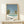 May include: Framed poster print leaning against white wall on light wood shelf. Shows poolside scene with turquoise swimming pool, two palm trees, clear blue sky, and white flowering bushes. Artist name and Tokyo 1973 text at bottom with colorful dots. Light wood frame matches shelf. Small wrapped gift box with greenery sits beside frame.