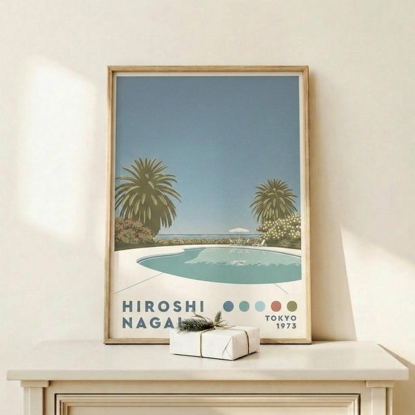 May include: Framed poster print leaning against white wall on light wood shelf. Shows poolside scene with turquoise swimming pool, two palm trees, clear blue sky, and white flowering bushes. Artist name and Tokyo 1973 text at bottom with colorful dots. Light wood frame matches shelf. Small wrapped gift box with greenery sits beside frame.
