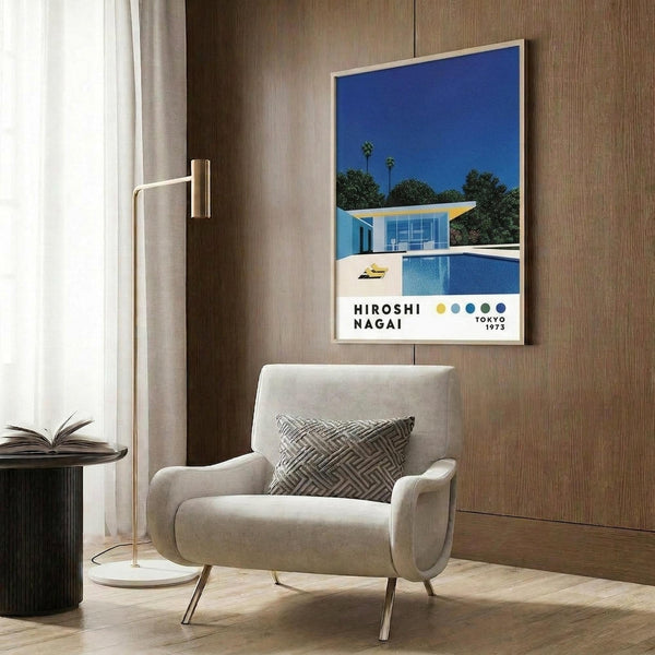 May include: Framed poster print displayed on wood-paneled wall showing modern blue house with swimming pool under bright blue sky. Print features color dots and Tokyo 1973 text. Scene includes beige upholstered armchair with geometric patterned pillow, gold floor lamp, dark round side table with open book, and light wood flooring in contemporary interior setting.