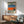 May include: Framed artwork displayed above wooden sideboard in modern living room with mid-century chair and table lamp. Print features woman with headphones against layered horizontal bands of pool water, orange surface, rocky ground, with two lounge chairs on deck above. Wood frame with light matting, natural sunlight streaming across white wall.
