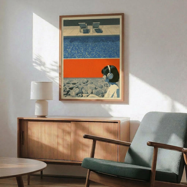 May include: Framed artwork displayed above wooden sideboard in modern living room with mid-century chair and table lamp. Print features woman with headphones against layered horizontal bands of pool water, orange surface, rocky ground, with two lounge chairs on deck above. Wood frame with light matting, natural sunlight streaming across white wall.