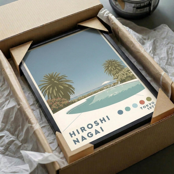 May include: Framed vintage-style poster showing palm trees beside a turquoise swimming pool against pale blue sky, with "HIROSHI NAGAI" and "TOKYO 1973" text and colorful dots. Black frame sits in cardboard shipping box with brown paper wrapping on white tissue paper.