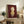May include: Framed poster artwork displayed on wooden sideboard in living room setting. Shows illustration of child figure with orange-red hair wearing yellow dress against deep burgundy background. Light wooden frame, positioned next to fern plant, books, and ceramic vessels. Warm neutral room with beige sofa visible.