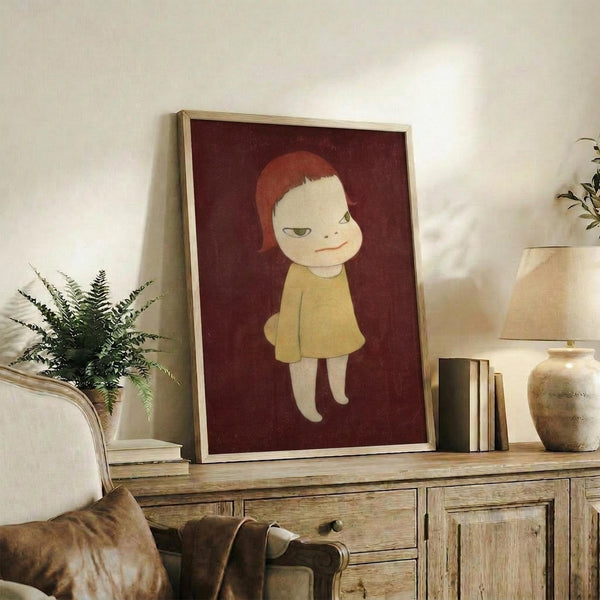May include: Framed poster artwork displayed on wooden sideboard in living room setting. Shows illustration of child figure with orange-red hair wearing yellow dress against deep burgundy background. Light wooden frame, positioned next to fern plant, books, and ceramic vessels. Warm neutral room with beige sofa visible.