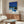 May include: Modern living room interior featuring a framed poster of a mid-century house with blue swimming pool against vibrant blue sky with palm trees, displayed above a walnut wood credenza. White curved chair and round glass coffee table in foreground, with white table lamp and copper floor lamp completing the minimalist Scandinavian-style decor.