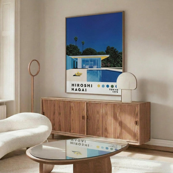 May include: Modern living room interior featuring a framed poster of a mid-century house with blue swimming pool against vibrant blue sky with palm trees, displayed above a walnut wood credenza. White curved chair and round glass coffee table in foreground, with white table lamp and copper floor lamp completing the minimalist Scandinavian-style decor.