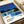 May include: Modern art print featuring a mid-century pool house with large glass windows, blue swimming pool, and palm trees against a bright blue sky. The minimalist illustration uses bold colors - blues, yellows, and whites - in a retro Japanese pop art style. Print sits on white paper against cream fabric background with soft lighting creating gentle shadows.