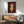 May include: Framed artwork showing a stylized child character with reddish-brown hair and pale skin wearing a yellow dress against a deep burgundy background, displayed above a mid-century modern wooden credenza with white table lamp, beside a sage green upholstered armchair with wooden frame in a sunlit room with white walls.