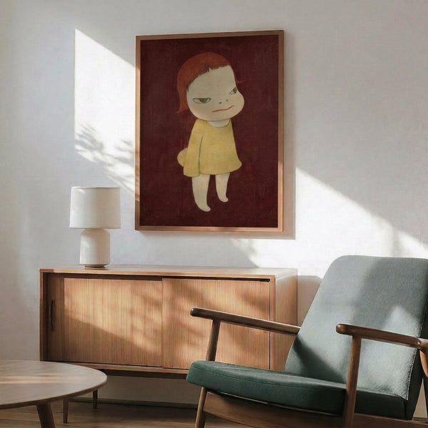 May include: Framed artwork showing a stylized child character with reddish-brown hair and pale skin wearing a yellow dress against a deep burgundy background, displayed above a mid-century modern wooden credenza with white table lamp, beside a sage green upholstered armchair with wooden frame in a sunlit room with white walls.