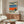 May include: Modern living room scene showing framed poster artwork on wall above wooden credenza. Artwork depicts poolside scene with blue water, orange stripe, rocky bottom section, and figure wearing white headphones. Mid-century modern furniture includes white curved chair, round glass coffee table, and walnut wood sideboard. Neutral beige walls with natural lighting and minimalist decor styling.